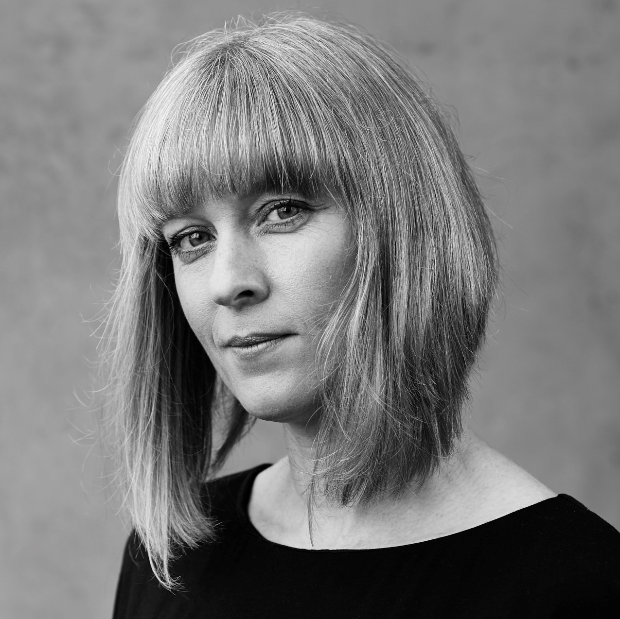 Black and white photograph of Sophie Woolley, a white woman, with long bobbed fair hair. She wears a plain black dress and looks brightly into the camera with a slight smile.