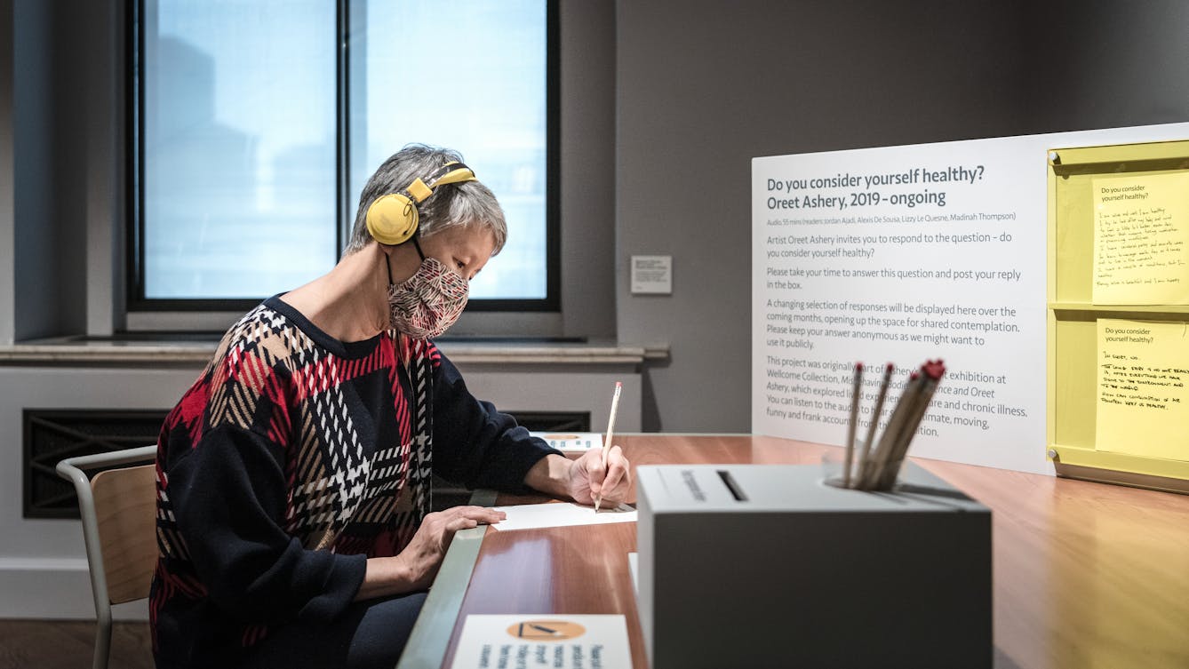 Photograph of an individual sat at a desk in a museum gallery setting, wearing a face covering and a pair of yellow on-ear headphones. They are holding a pencil in their left hand, writing on a sheet of paper. To their right is a small post box and a pencil holder. Behind them is a window. In front of them is a large information panel with the title, 'Do you consider yourself healthy? Oreet Ashrey, 2019 - ongoing' written on it.