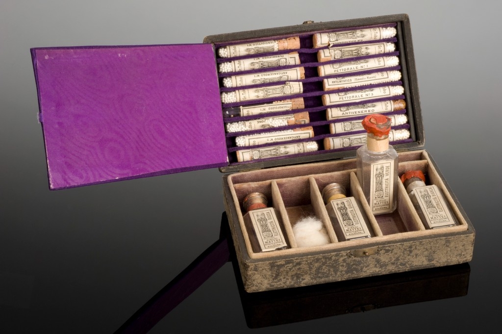 Photograph of open box containing medicine in vials and bottles, and one ball of cotton wool.  One bottle has been stood up vertically for the photograph.  The inside of the box's lid is covered with purple material.