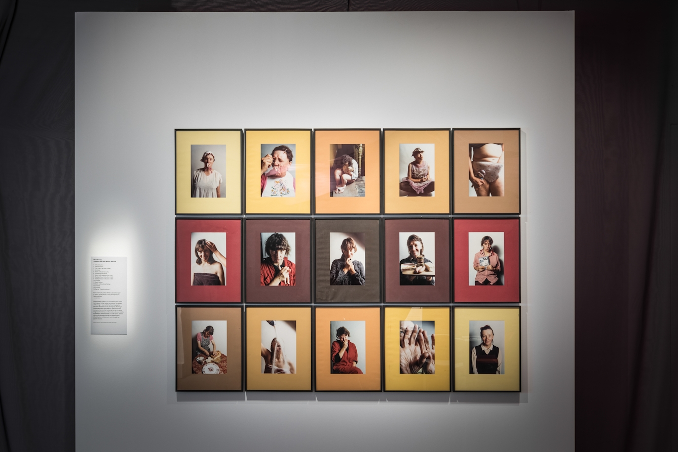 Photograph of a grid of framed photographs. The images show a woman in various self portrait scenarios. The photographs are framed in mounts in the hues of yellows, oranges and browns.