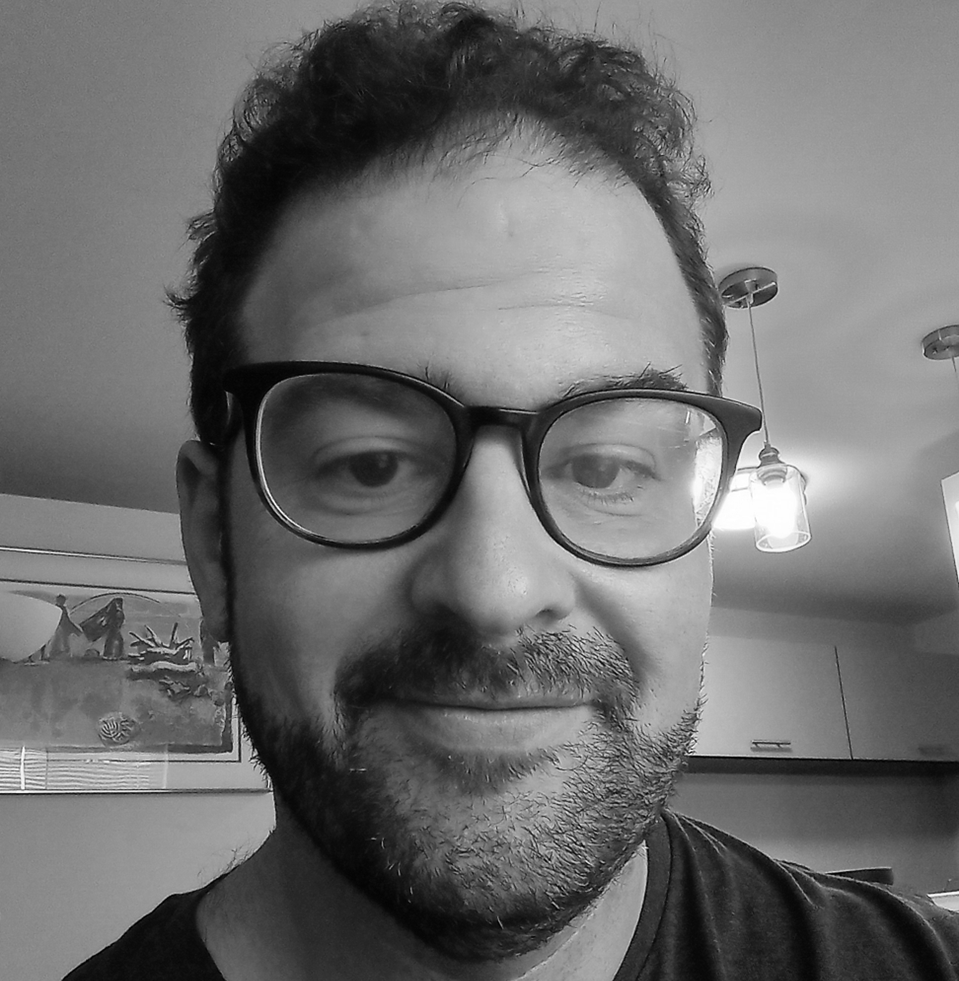 Black and white photograph of the head of a young white man with short dark hair and large black-framed glasses. He is smiling and looking into the camera.