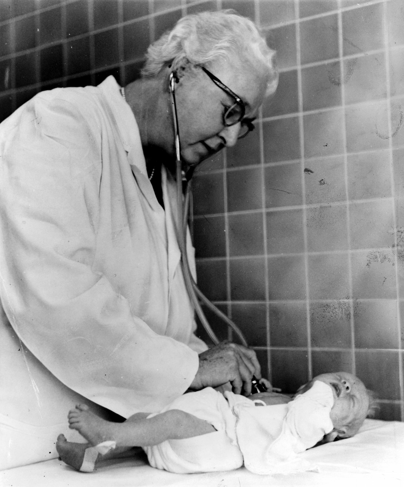 A black and white image of Virginia Apgar, in a white medical coat, standing over a newborn baby and using a stethoscope to measure their heart rate.