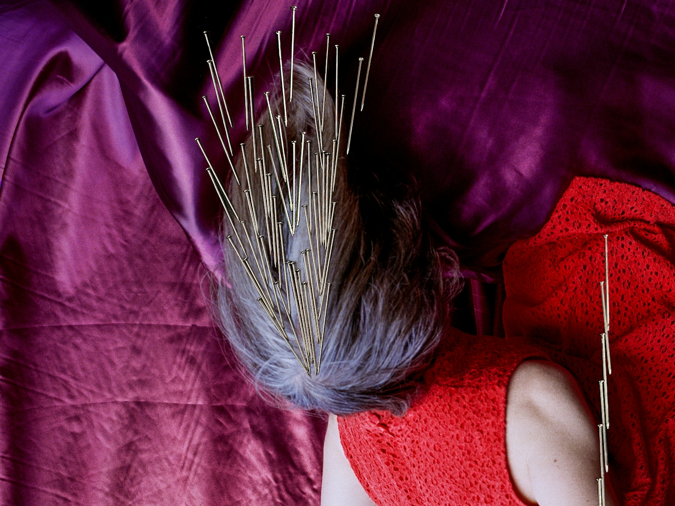 Detail from a larger artwork created with a colour photographic print of a female figure in a bright red dress, set against a purple and blue draped silk background. She is curled up in the frame, caught as if in mid fall, hair blown upwards. Her face is obscured by her hair. Her body is targeted by groups of dress pins, laid on top of the photographic print. The pins are arranged as if they are a flight of arrows directed at her body. One group descends from above towards the crown of her head.