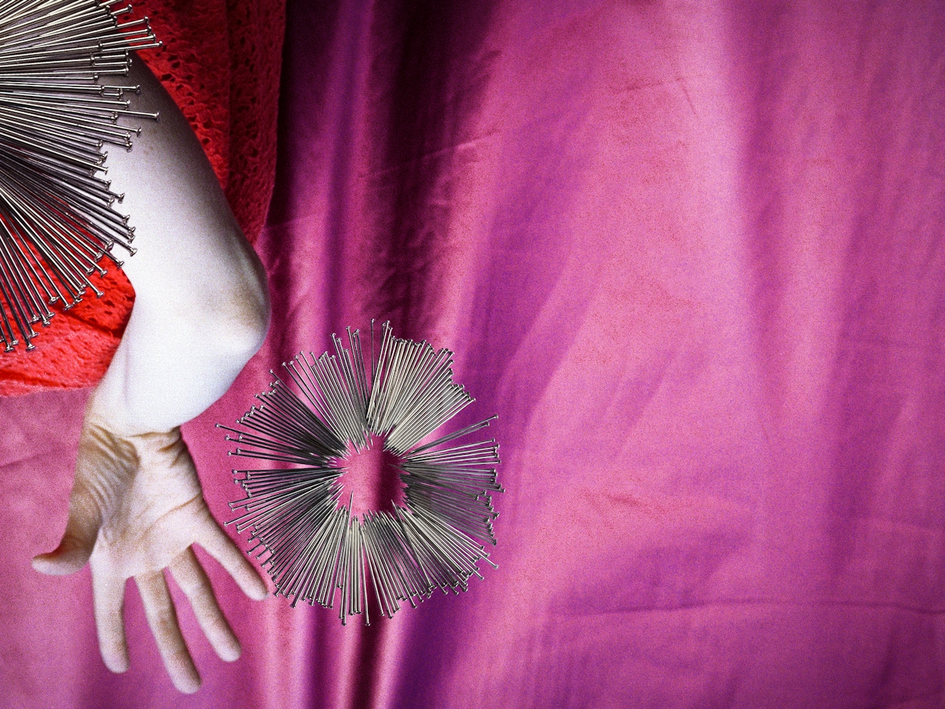 Detail from a larger artwork created with a colour photographic print of a female figure in a bright red dress, set against a purple and blue draped silk background. The figure is on her side, her arms are extended out behind her back, bent at the elbow, with the fingers splayed out. Her body is covered by groups of dress pins, laid on top of the photographic print. The pins are arranged in a circle so that the sharp end of the pins point to the centre of the circle and float just to the right of her outstretched hand.