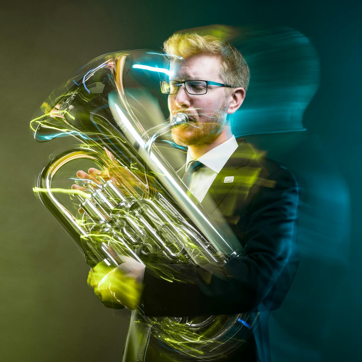 Photograph of a young man playing a euphonium against a dark background. The slow exposure of the camera causes his movements to be traced across the image as a blur. He is surrounded by wisps of smoke which are coloured yellow on the left of the frame and a blue on the right.