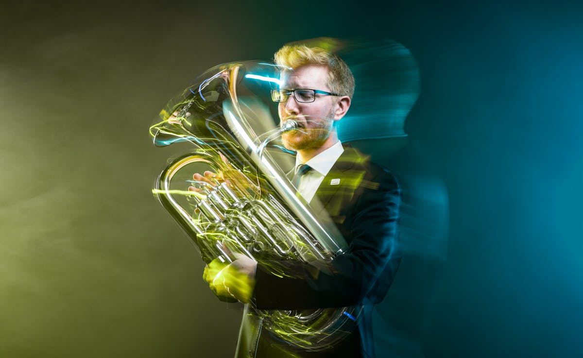 Photograph of a young man playing a euphonium against a dark background. The slow exposure of the camera causes his movements to be traced across the image as a blur. He is surrounded by wisps of smoke which are coloured yellow on the left of the frame and a blue on the right.