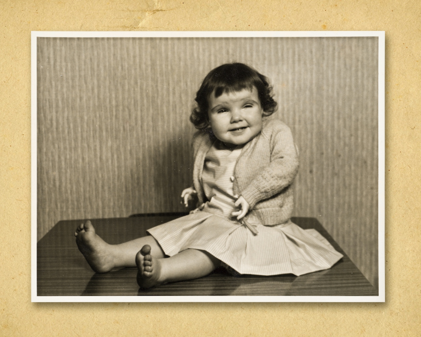 Photograph of a black and white photographic print, resting on a brown paper textured background. The print shows a small young girl sitting with her legs stretched out in front of her, on what looks like a tabletop. Behind her is a wall covered in a textured wallpaper with a vertical pattern. She is wearing a dress and cardigan. She is smiling and looking off slightly right of camera. The arms in her cardigan sleeves are prosthetic arms. The girls mother was prescribed Thalidomide during her pregnancy.
