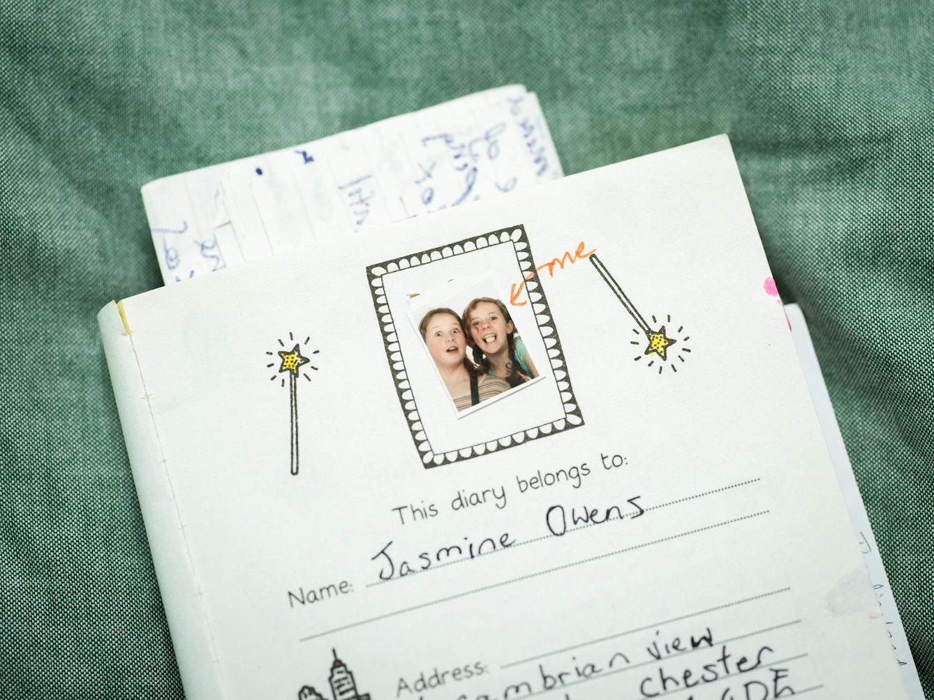 Photograph of the top half of the first page in a young child's paper diary. At the top of the page is a photo booth print of two young girls making silly faces to the camera. There is a hand drawn red arrow and the word 'me' pointing to one of the girls. Beside the photo are 2 illustrated wands with yellow stars on the top. Under the photo is written, 'This diary belongs to, Name: Jasmine Owens. Address....'. The diary has loose pages protruding from the top and is lying on a green coloured fabric background.