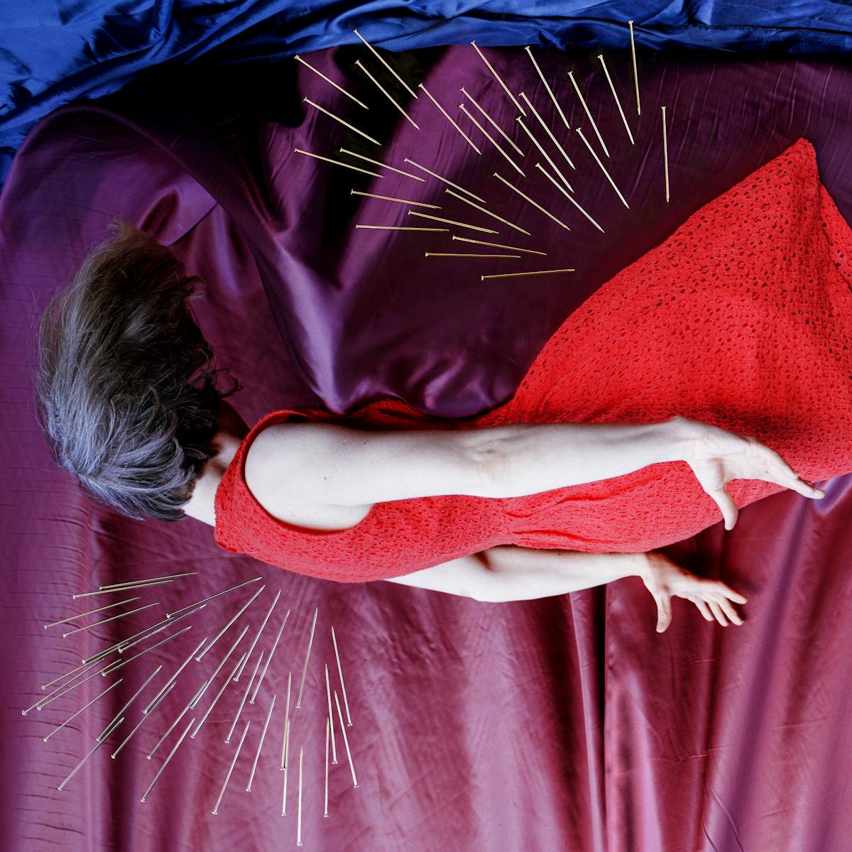 Artwork created with a colour photographic print of a female figure in a bright red dress, set against a purple and blue draped silk background. She is lying horizontal in the frame, caught as if in mid fall, hair and dress blown upwards, toes and fingers outstretched. Her face is obscured by her hair. Her body is surrounded by groups of dress pins, laid on top of the photographic print. The pins are arranged as if they are a flight of arrows directed at her body. One group attacks her back from underneath, another her feet and ankles from above and below. A final group descend from above towards her billowing skirt.