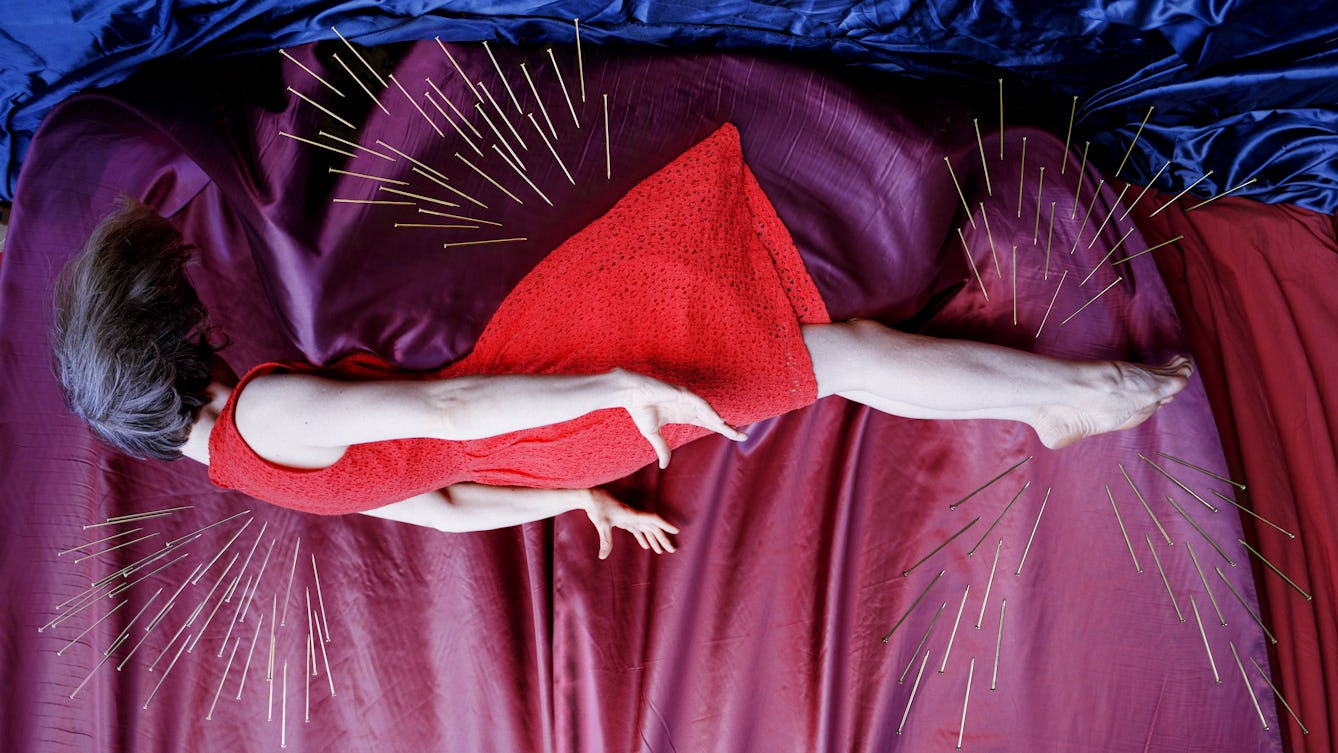 Artwork created with a colour photographic print of a female figure in a bright red dress, set against a purple and blue draped silk background. She is lying horizontal in the frame, caught as if in mid fall, hair and dress blown upwards, toes and fingers outstretched. Her face is obscured by her hair. Her body is surrounded by groups of dress pins, laid on top of the photographic print. The pins are arranged as if they are a flight of arrows directed at her body. One group attacks her back from underneath, another her feet and ankles from above and below. A final group descend from above towards her billowing skirt.