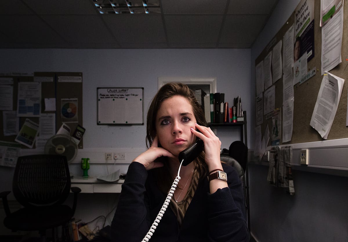 Photograph of a woman sat in an office environment. She is holding a landline telephone receiver to her left ear and is looking into the distance over the top of the camera.