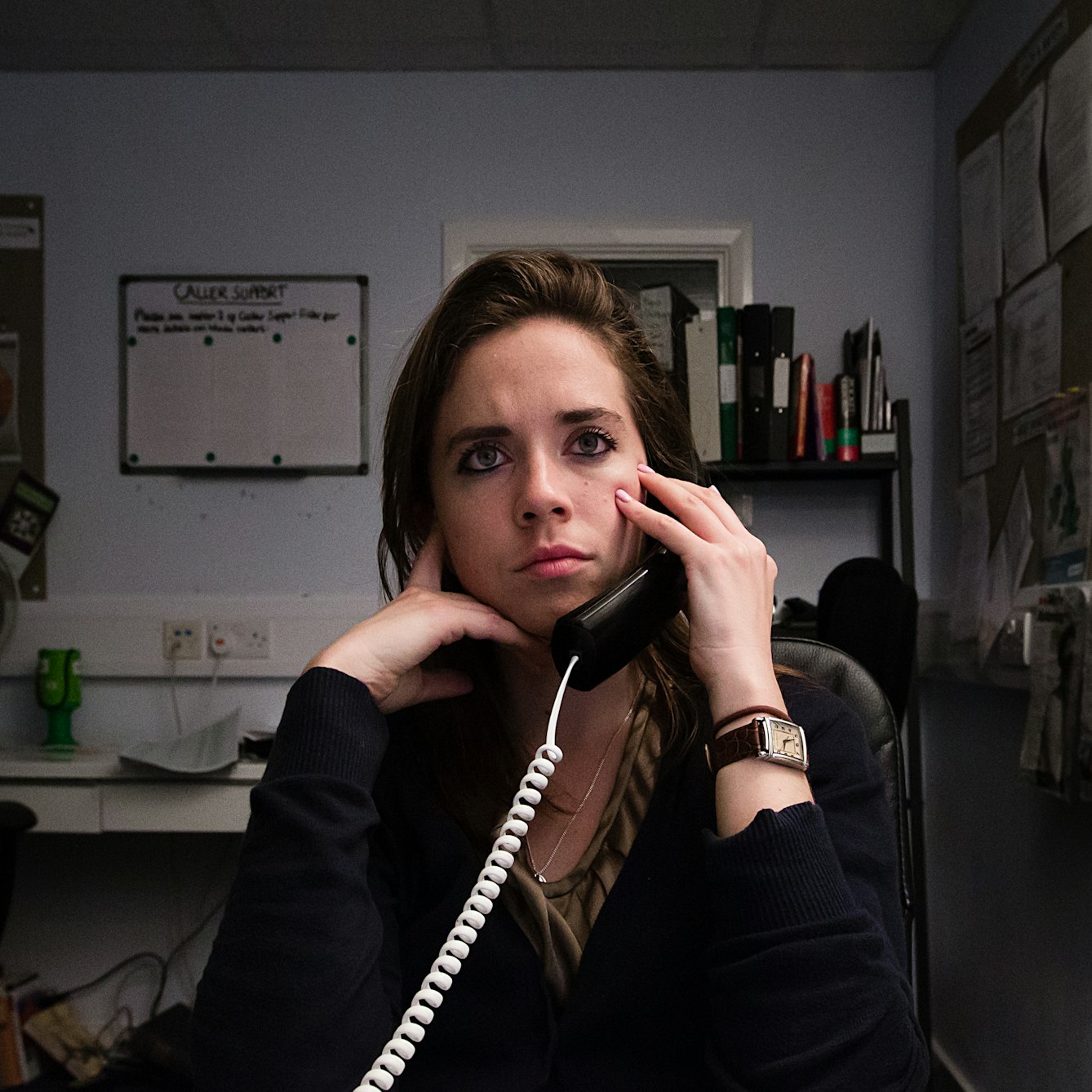 Photograph of a woman sat in an office environment. She is holding a landline telephone receiver to her left ear and is looking into the distance over the top of the camera.