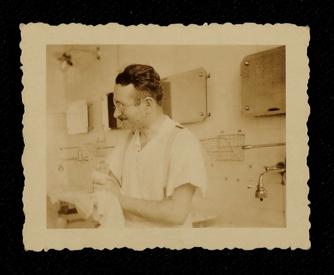 Sepia photographic print of Ludwig Guttmann in his medical practice in the 1930s. Guttmann has wavy dark hair, glasses and a moustache just above his top lip. He is wearing a white, short-sleeved shirt and braces. He is wiping his hands on a light coloured cloth and appears to be smiling and looking away to the left of the image. Behind Guttman are lots of wall-mounted mirrors, taps and baskets on a tiled wall.