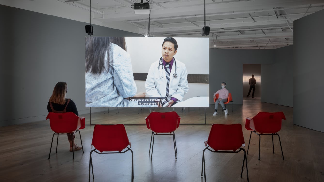 Photograph of a gallery space with grey walls and a wooden floor. Hung from the ceiling is a large screen, onto which a video is projected. the video shows a man in a white coat with a stethoscope around his neck talking to someone whose back can be seen to the left of the video. Subtitles can be seen on the screen. In the foreground and background are red plastic chairs. One person is sitting facing the screen, another is sitting facing the camera. Both are wearing face coverings.
