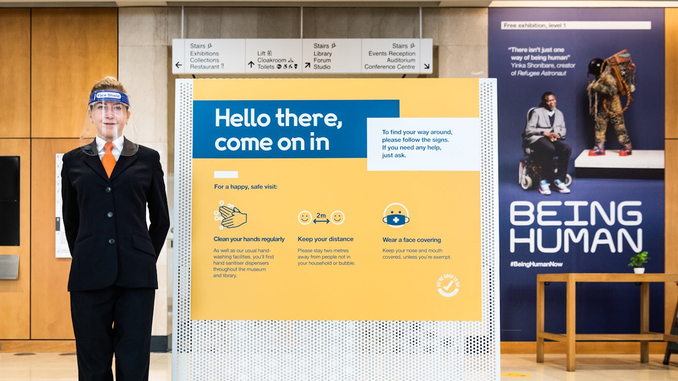 Photograph of the entrance to a museum showing a large poster with the text, 'Hello there, come on in' written in large letters. To the left of the sign is a smiling female security guard wearing a transparent visor on her head. To the right of the sign in the distance is a large banner on the wall showing a photograph of a man seated in a wheelchair next to a large mannequin artwork. Under the photograph are the words, 'Being Human'. 
