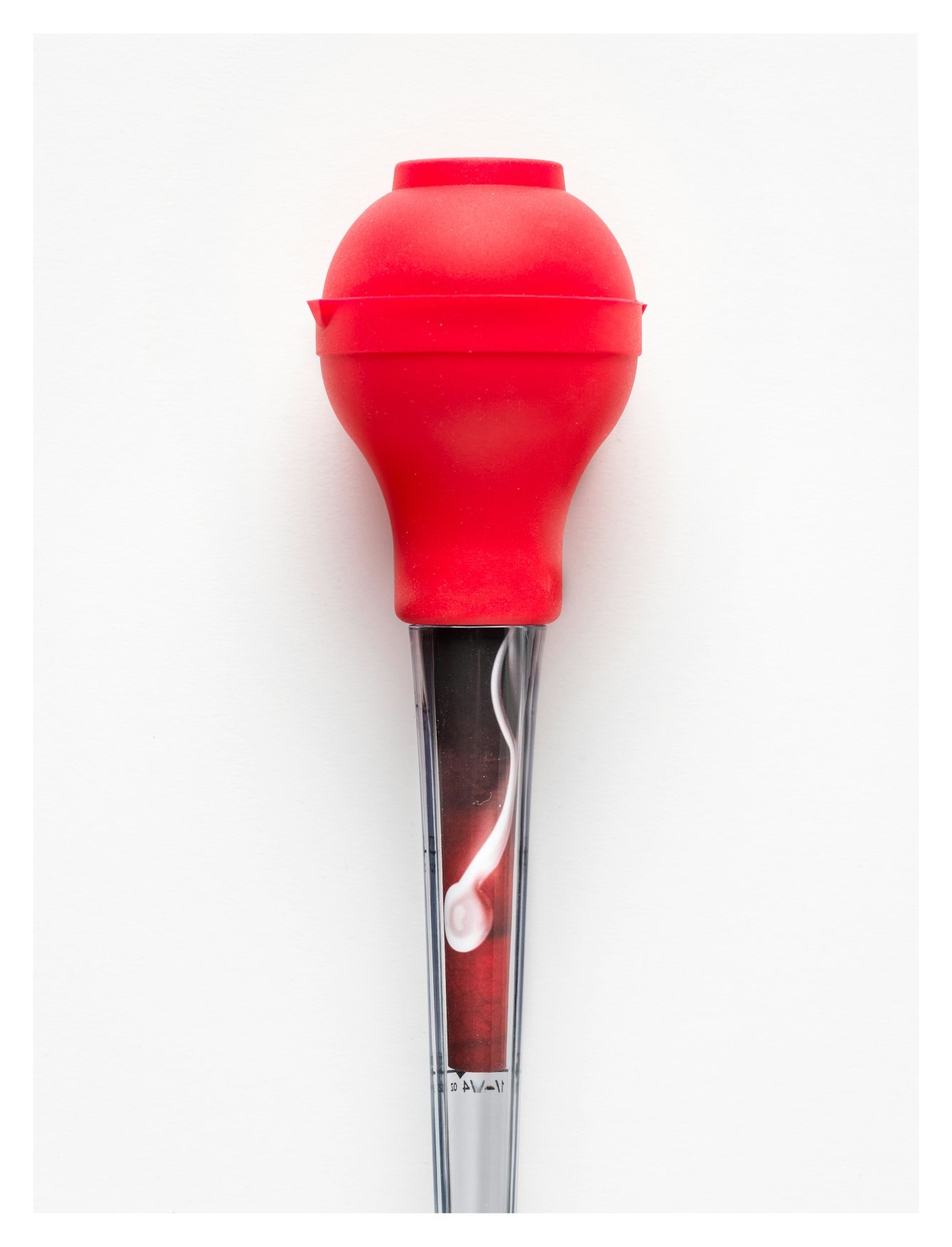 Photograph of a glass turkey-baster with a bright red rubber bulb, against a white background. Rolled up in the glass tube is a colour image of a single white sperm against a red background.