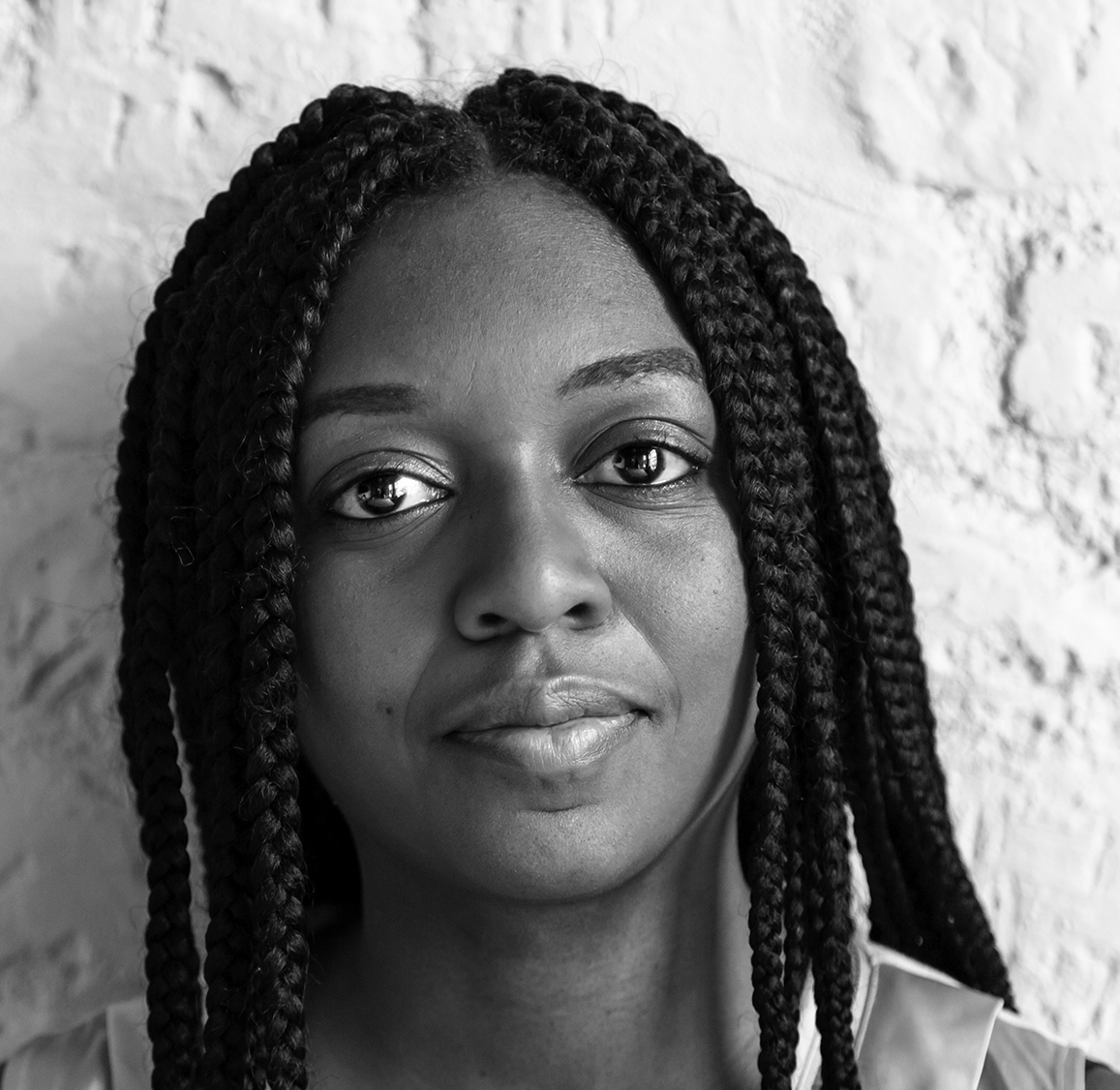 Balck and white photograph of the head and shoulders a young black woman with long braided hair. She is looking directly at the camera.