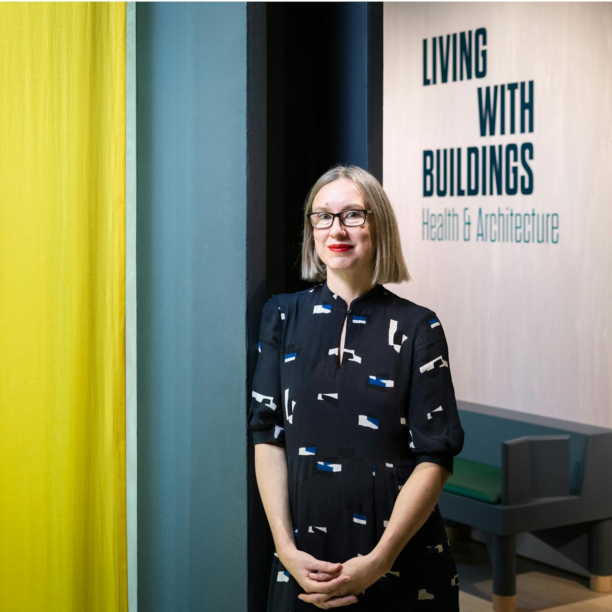 Photographic portrait of Emily Sargent, curator of the exhibition Living with Buildings at Wellcome Collection. Emily is leaning against a pillar within the exhibition space, with a hanging yellow drape to the left and the title of the exhibition to the right.