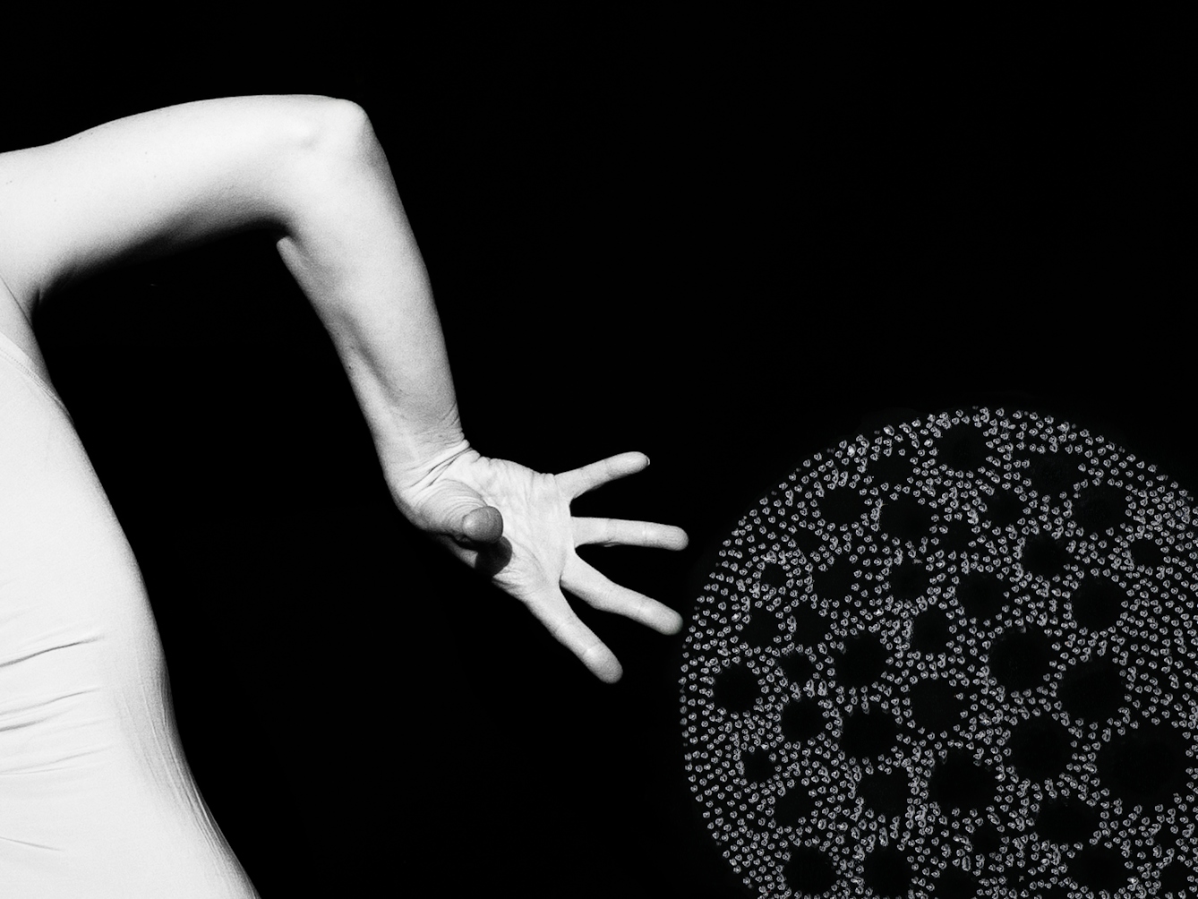 Artwork made up of a black and white photograph of part of a female figure from behind, from the waist up, against a black background. Her body is twisted in a dancerly pose.  Her right arm is bent down to the ground, hand outstretched. To the right and below her right hand is another large circular shape with holes in the middle, made up of a layered texture of grey dots which are painted onto the print creating a three dimensional texture.