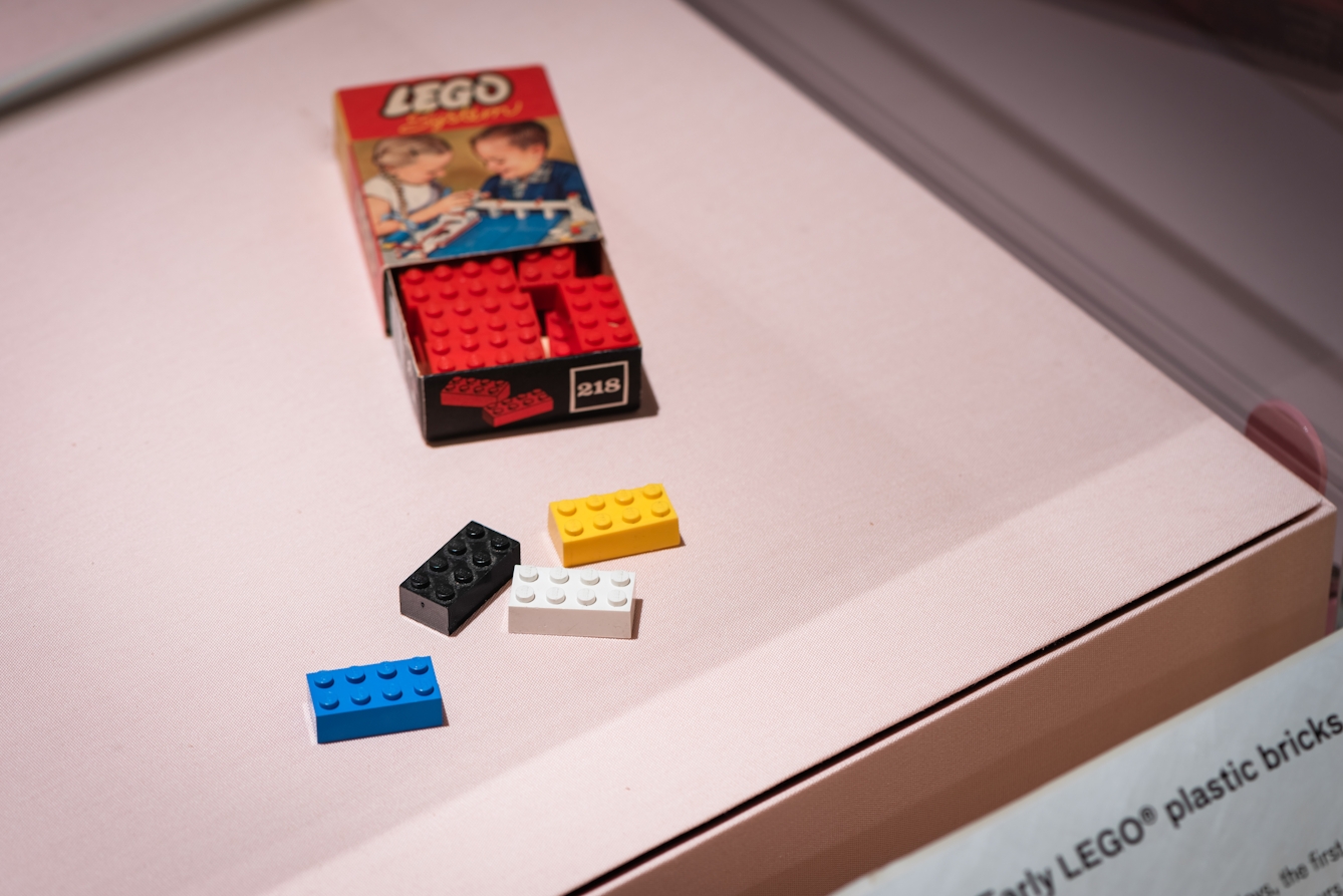 Photograph of an exhibition display case containing an early LEGO packaging box half open, containing red bricks. In front of the box are 4 LEGO bricks, coloured black, yellow, blue and white.