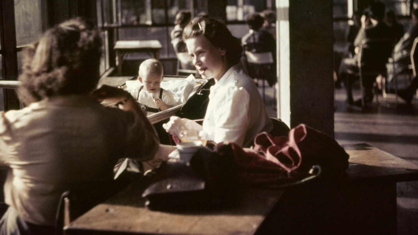 Photograph from the mid-20th Century Peckham Pioneer Centre, showing two women seated at a table, speaking, whilst a baby plays in a pram behind them and in the distance another group sit at tables. The building in which they sit has concrete columns and large windows.