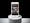 Photograph of a white mobile phone on a grey cylinder, with a grey and black background. On the phone is a twitter page displaying a composite photograph of Bernie Sanders sitting in front of a woman in a red dress, surrounded by a watching crowd. The twitter text reads “Performance Art specifically allows for emotional as well as multi sensory experiences rather than just intellectual ones.