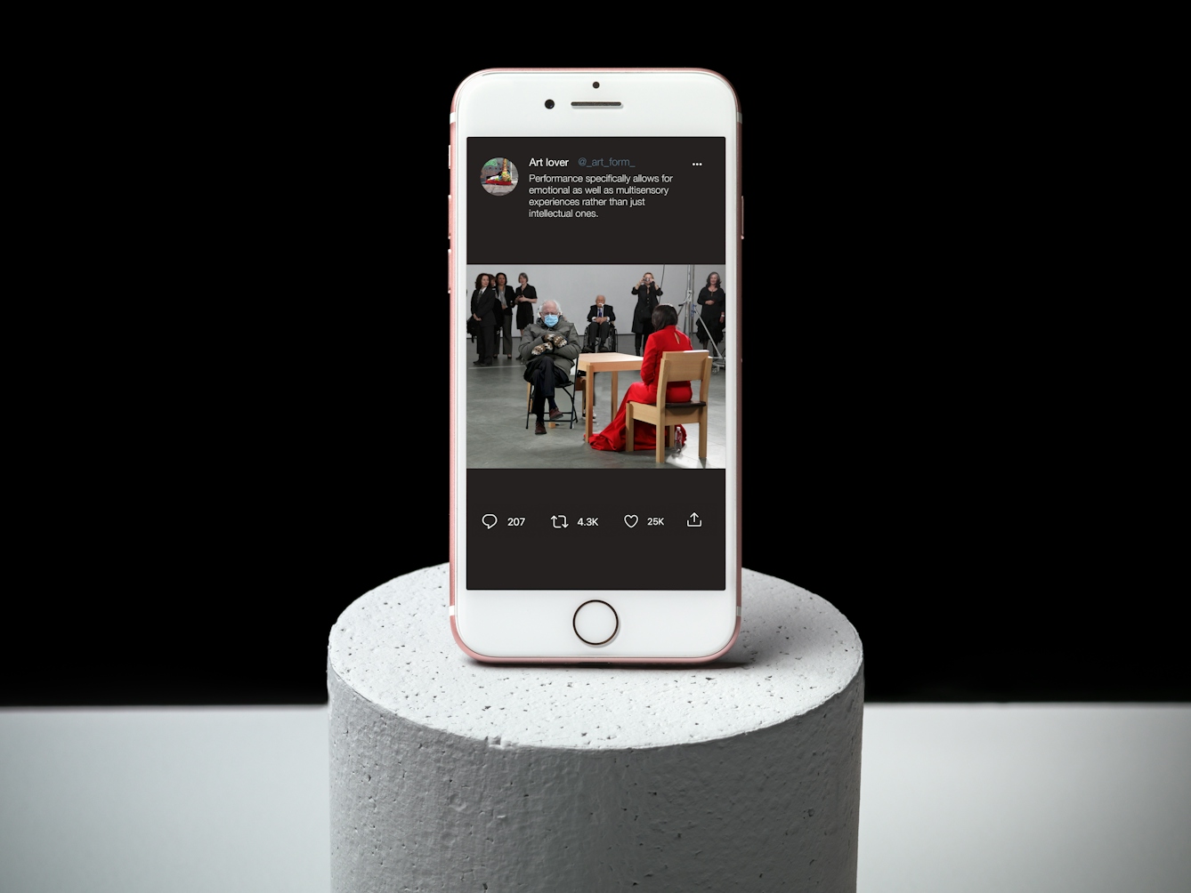 Photograph of a white mobile phone on a grey cylinder, with a grey and black background.  On the phone is a twitter page displaying a composite photograph of Bernie Sanders sitting in front of a woman in a red dress, surrounded by a watching crowd.  The twitter text reads “Performance Art specifically allows for emotional as well as multi sensory experiences rather than just intellectual ones.