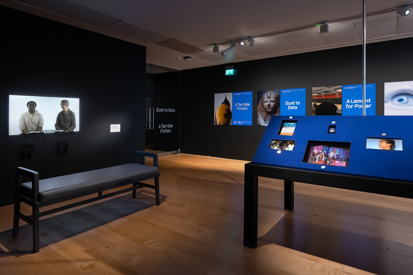 Photograph of an exhibition gallery space made up of black painted walls and a wooden floor. To the left of the scene is a video installation showing two individuals in conversation. In front of the screen is a long backless bench. To the right of the scene is a display case containing mounted screens showing video content set into a blue fabric backboard. In the distance, mounted on the wall are several large photographs accompanied by titles. There's a photograph of a taxidermy bird with the title A Terrible Fiction, a photograph of a sphinx with the title Dust to Data and a computer generated image which is partially obscured with the title A Lament for Power.