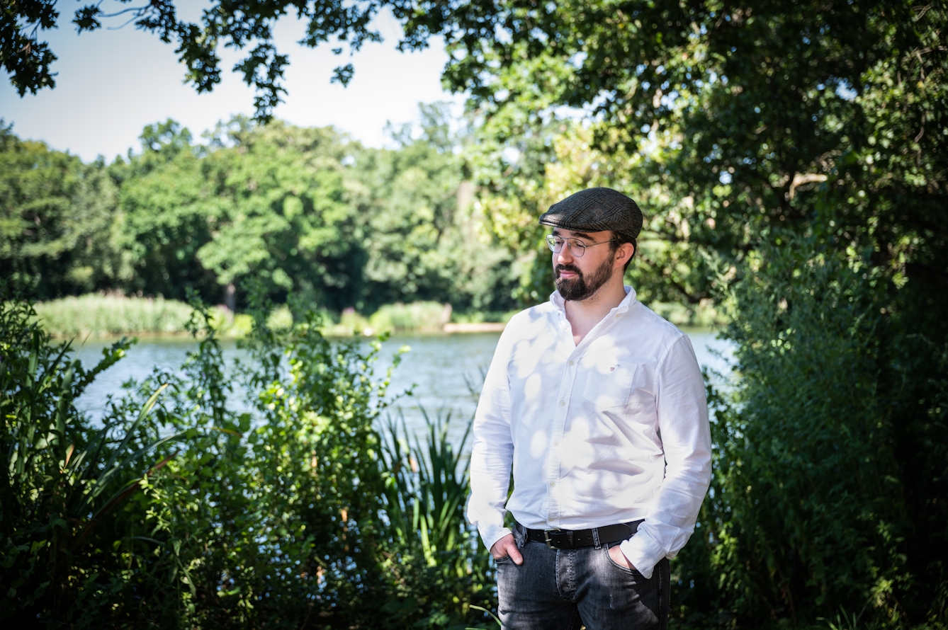 Photograph of a man from the waist up standing outside in a park like environment. He is wearing a flat cap, a white shirt and black jeans. He is looking towards the ground to camera left with a neutral expression. His hands are sunk into his pockets. Around him are lush green foliage from shrubs and trees. In the middle distance is an expanse of water and then in the far distance is the shoreline on the other side, with more grasses and trees. He is framed by the leaves of an overhanging branch beyond which is a small patch of blue sky.
