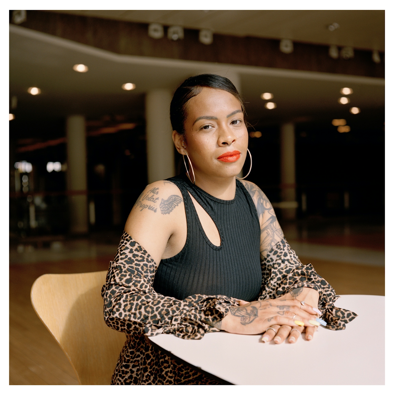 Portrait photograph of Lauren, a young Black woman, sitting on wooden chair. She has several tattoos on her arms and is wearing a black sleeveless top and a leopard print skirt. She has large hoop earrings and is wearing an orange red lipstick. 

She is looking straight to camera with a quiet smile. Behind her, there is an exhibition space which is empty apart from several wooden chairs. 