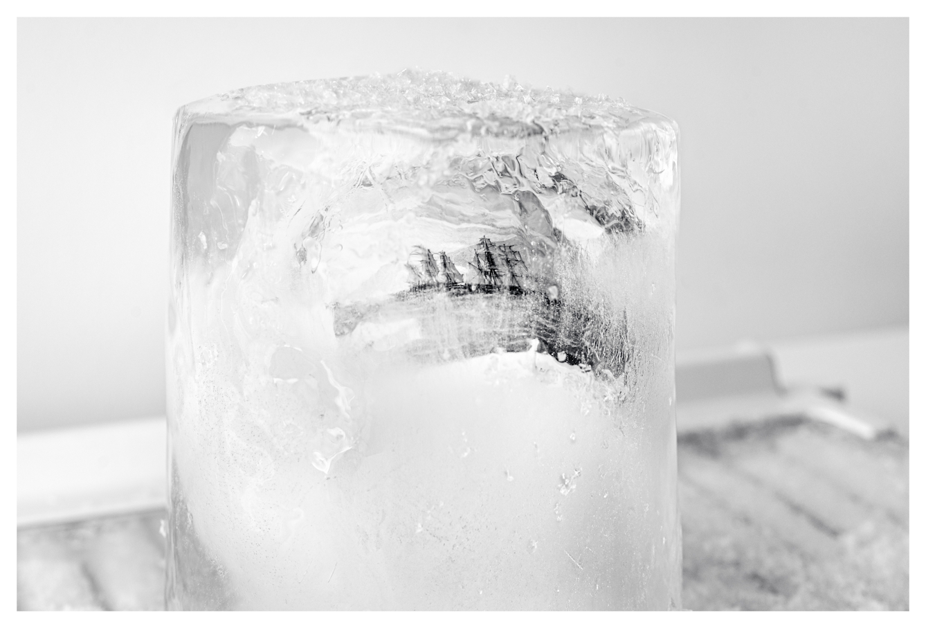 Monotone photograph showing a cylindrical core of frozen water. Encased within the ice is an illustration of tall sail ships from the 1800s that can just be seen through the distortions of the ice wall and opaque frosting. The ice core is standing vertically on a fridge freezer shelf.