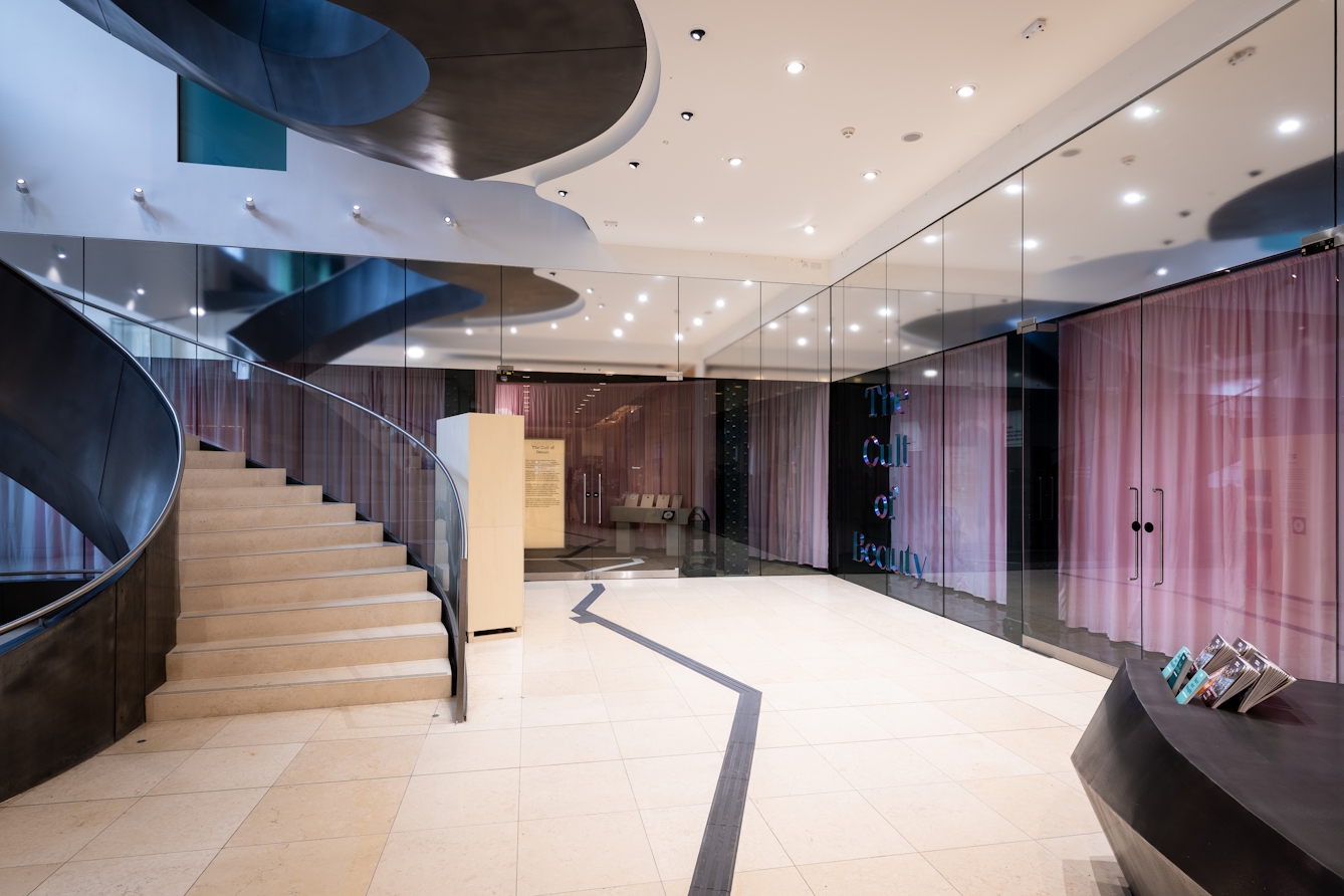 Entrance to Gallery 1, Wellcome Collection, London. A modern sweeping staircase is on the left. The glass walls and doors of the gallery are at the back of the lobby area, the information desk is in the foreground on the right.