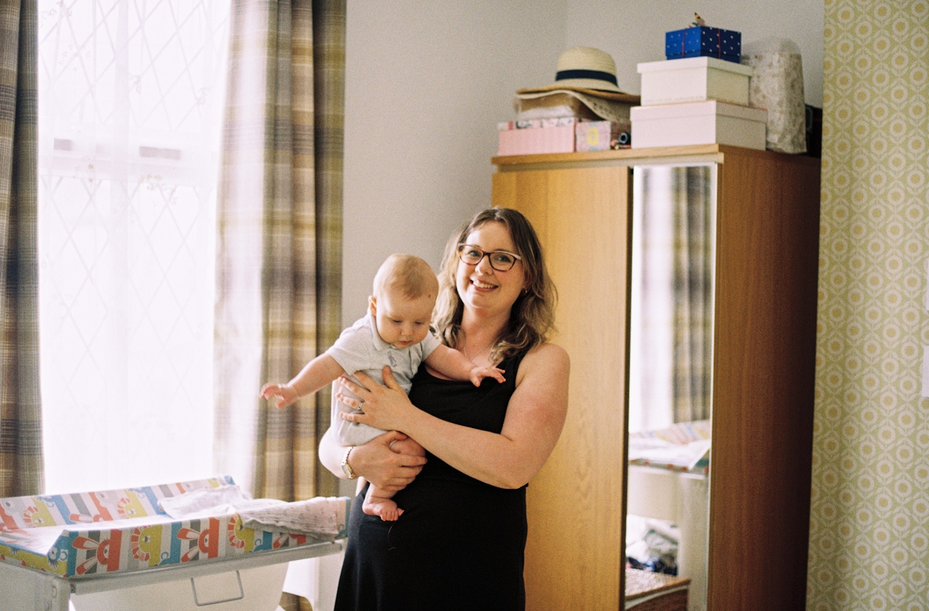 Photograph of a mother holding her baby son taken in a bedroom setting. The mother is looking to camera smiling. The baby has his arms outstretched looking down to the ground. Behind them is a large window through which light is flooding in. Also in the background is a tall wardrobe with a mirror and stacks of boxes and summer hats on top, next to a patterned wallpapered wall. Beside them is a cot with a changing mat resting on top is just visible.