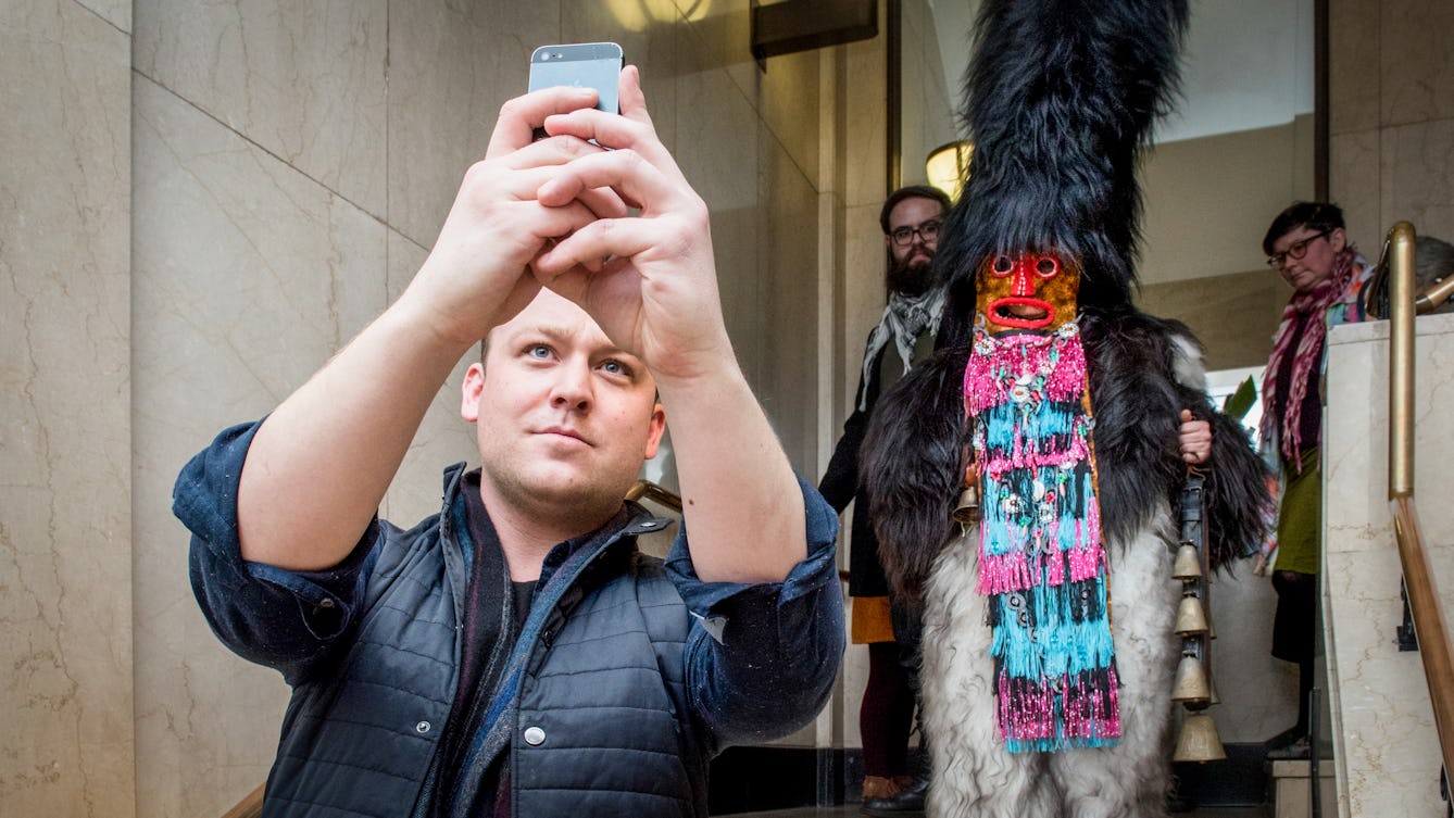 Photograph at a late night museum event. On the left hand side a man holds up a mobile phone in both hands up in front of his head, taking a photo over his shoulder of the scene behind him. The scene his is photographing is a person descending a staircase dressing in a fur outfit complete with mask and tall fur covered headdress. They are carrying a set of bells.