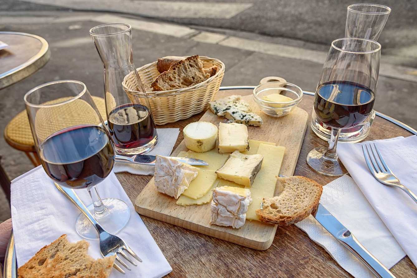 Photograph of a small table laid out with cheese, wine, bread, napkins and cutlery.