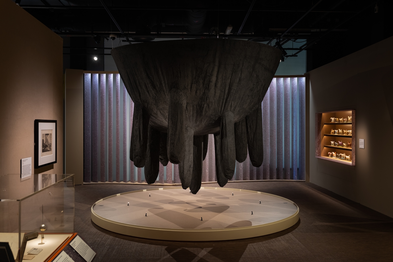 Photograph of a large art installation set within a larger exhibition gallery space. The installation resembles a large black udder-like sculptural form, suspended from the ceiling. Surrounding the sculpture are other exhibits, framed works on the wall and exhibition cases.