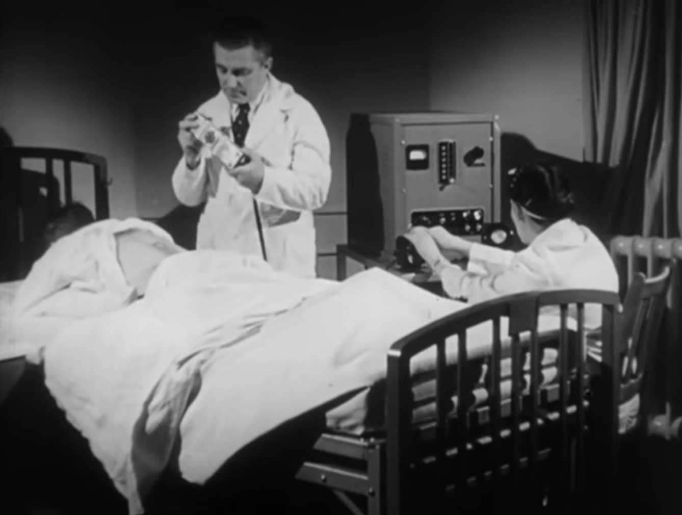 Image of a patient lying on his stomach in bed with his shirt lifted to expose his back. A man wearing a lab coat stands over him and a woman wearing a lab coat operates a box-like machine next to the bed.