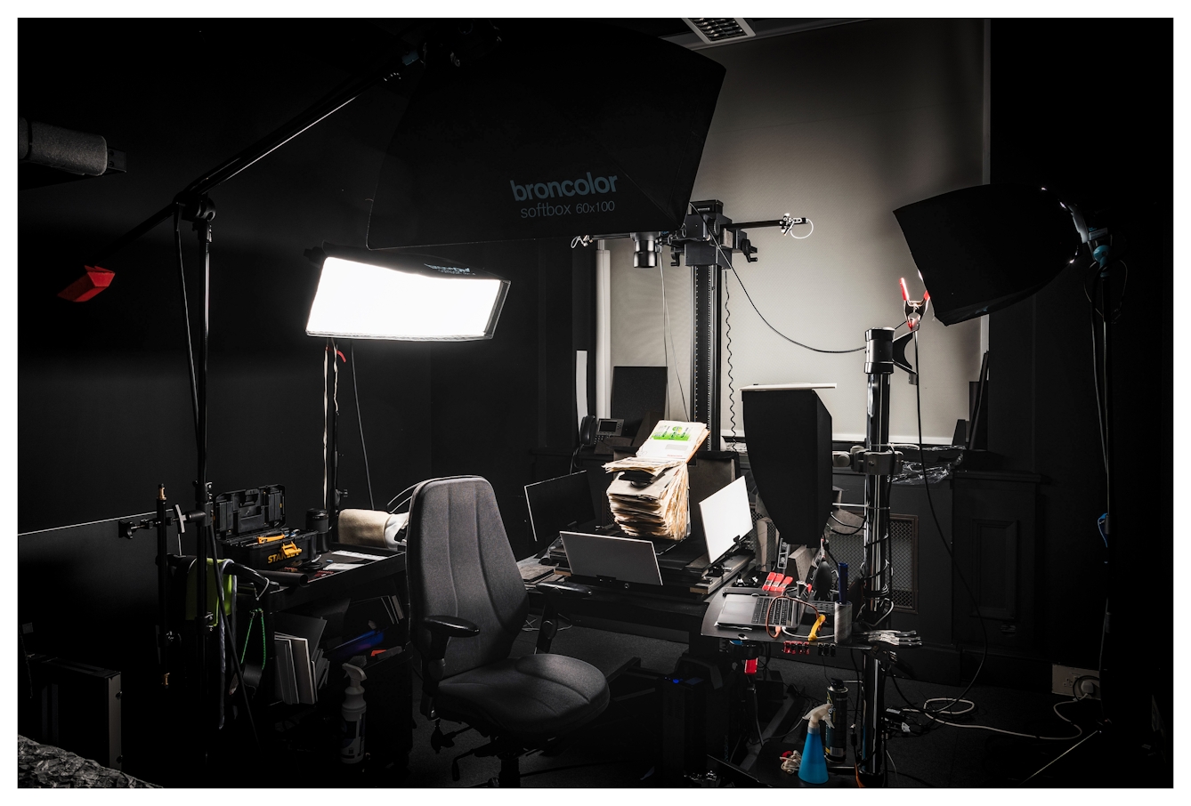 Wide angle photograph of a dark photographic studio. In the centre of the image is an open scrapbook positioned on a table underneath a camera which is mounted vertically above it. Around the scrapbook is a trolley with a laptop and a display, 3 large studio lights and another trolley containing various accessories. The room has an atmospheric feel, all dark except for a pool of light in the centre, spotlighting the scrapbook.