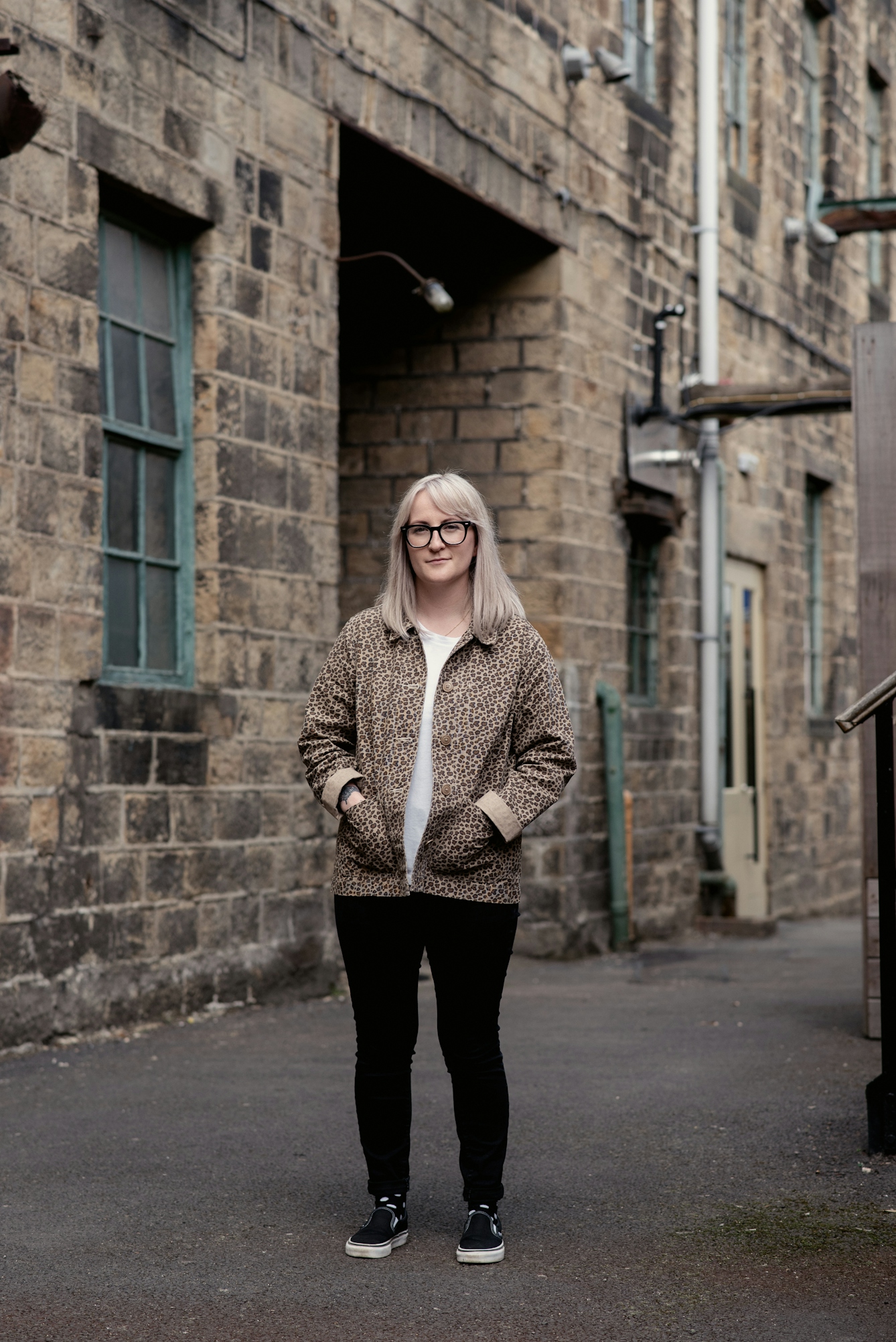 Photograph of a young woman with shoulder length blond hair and glasses in full length. She is standing in an industrial back-street location, wearing a white t-shirt, a leopard skin patterned jacket and black leggings. Her hands are in her jacket pockets and she is looking to camera with a slight smile. Behind her in the background is a large brick building with windows and doorways and external pipework and cables.