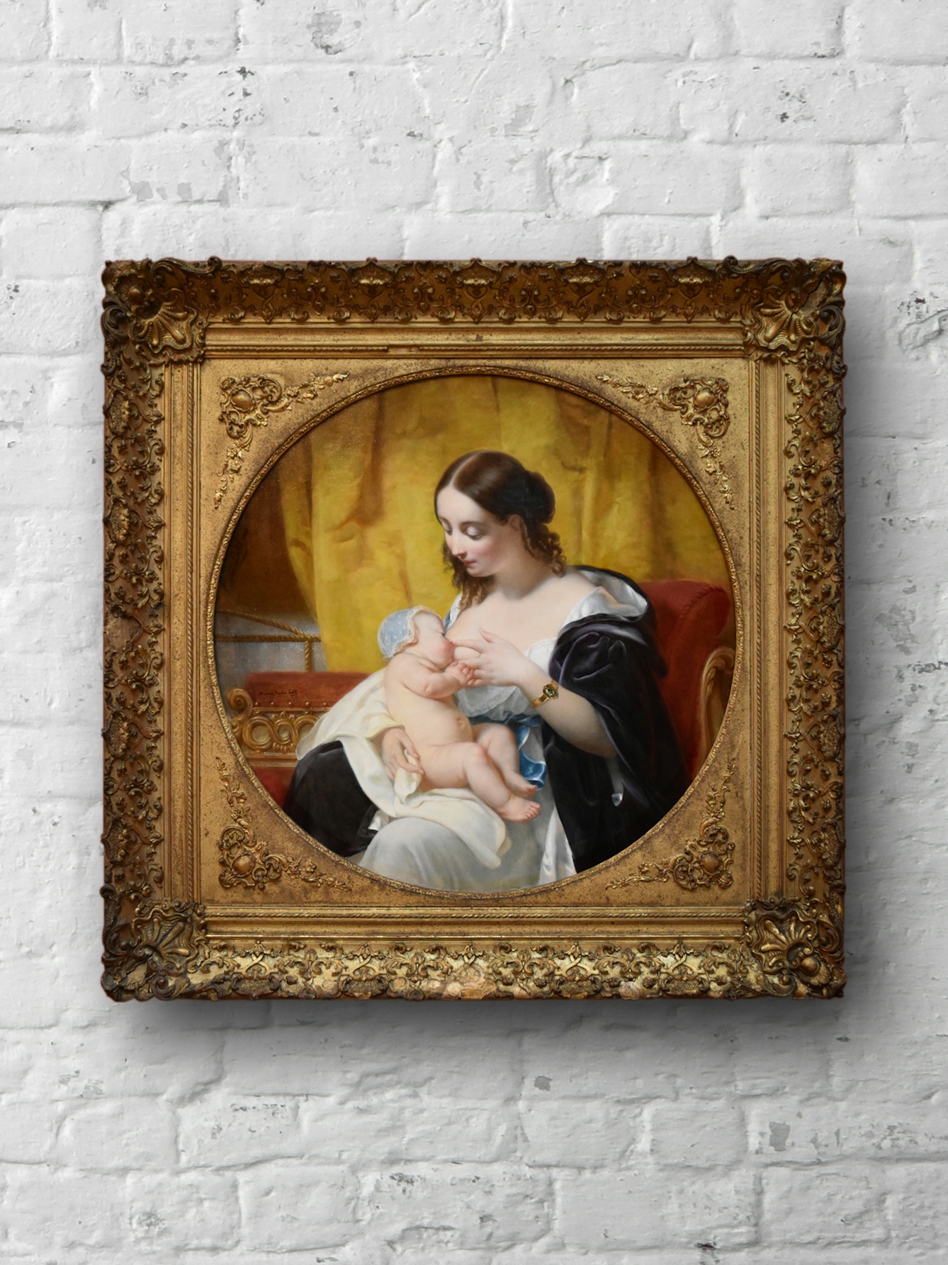 An image of an oil painting depicting a women acting as a wet nurse and breastfeeding a baby. The women is shown cradling the small child with one arm and holding her breast to the child's mouth. The painting is in an ornate gilt frame with a circular aperture in the middle, and set against a white brick background.
