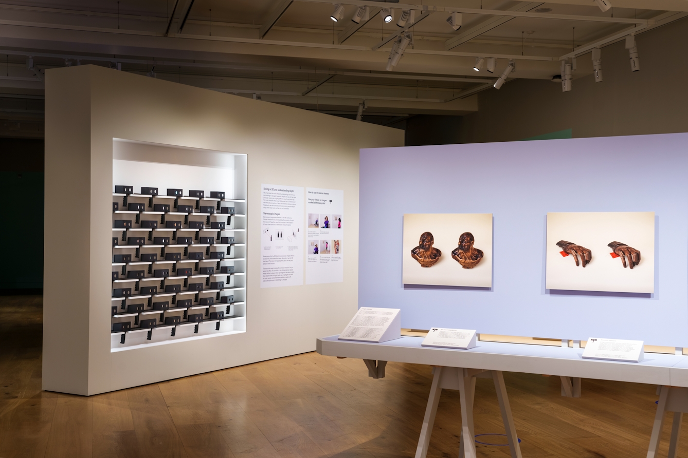 Photograph of a gallery exhibition space showing large photographic prints mounted on the walls and free standing display boards. The colours of the room are light whites and purples. On one of the walls is a rack of shelving which contains many stereoscopic viewers, ready for visitors to take and use.