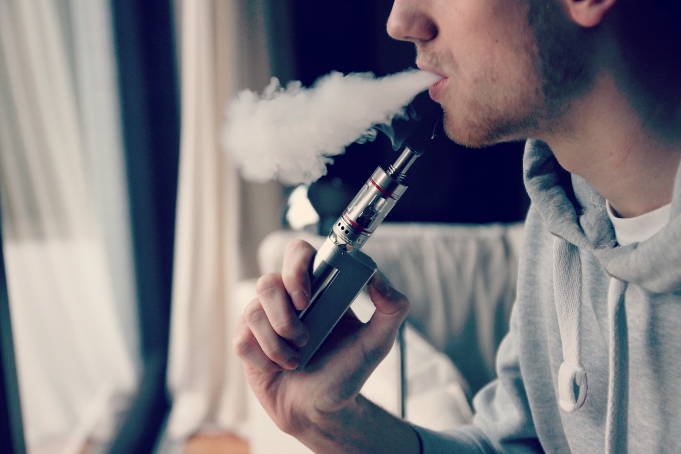 Colour photograph showing a white man in a grey hoodie holding a vaping device close to his mouth and exhaling smoke.