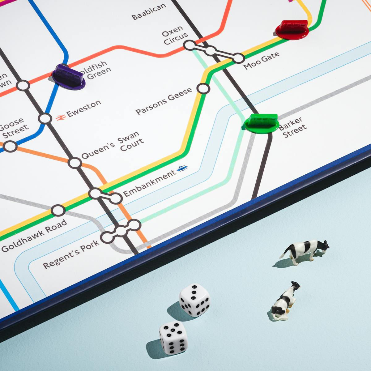 A photograph of a bottom right section of a board game featuring a city underground map with animal place names such as Moo Gate, Ham Heath and Oxen Circus is visible in the top left of the frame. In the bottom section are two die with the numbers five and three, 5 toy cows and a toy London Underground Carriage.