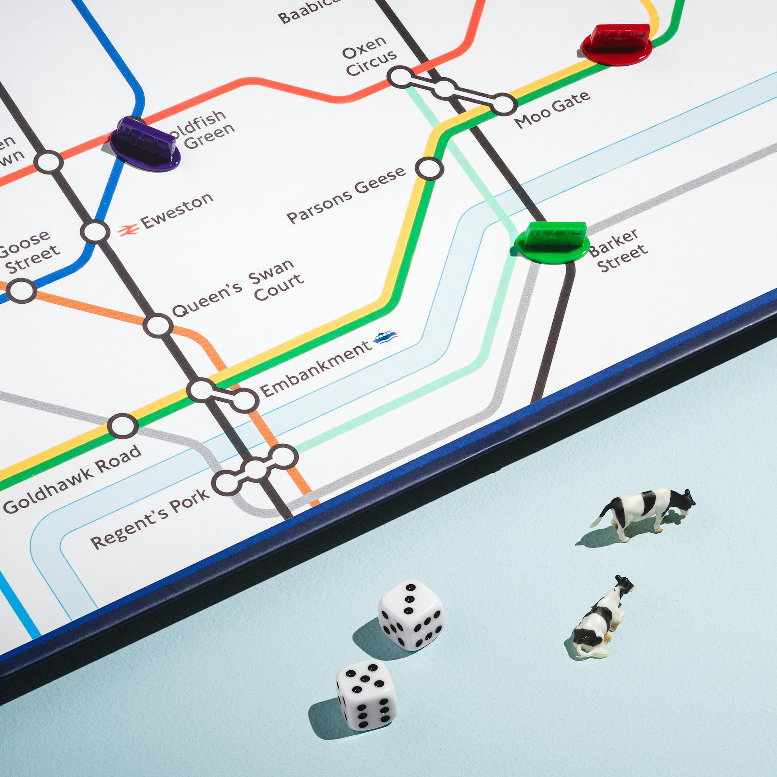A photograph of a bottom right section of a board game featuring a city underground map with animal place names such as Moo Gate, Ham Heath and Oxen Circus is visible in the top left of the frame. In the bottom section are two die with the numbers five and three, 5 toy cows and a toy London Underground Carriage.