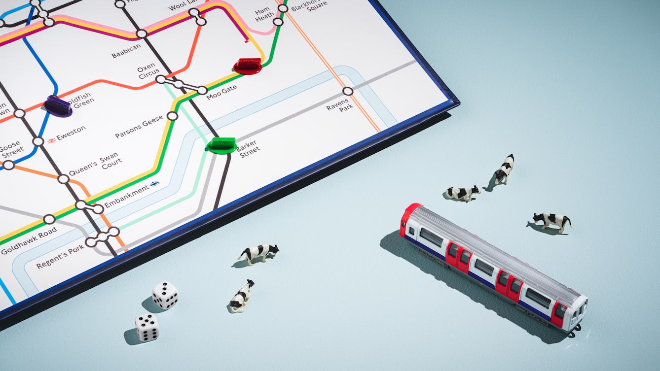 A photograph of a bottom right section of a board game featuring a city underground map with animal place names such as Moo Gate, Ham Heath and Oxen Circus is visible in the top left of the frame. In the bottom section are two die with the numbers five and three, 5 toy cows and a toy London Underground Carriage.