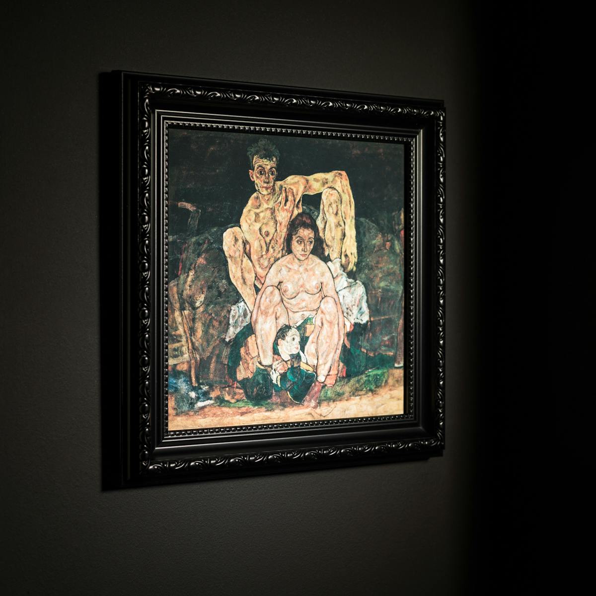 Photograph black ornate picture frame hung on a black wall. Inside the frame is a print of an oil painting by Egon Schiele titled, 'The Family'. It depicts  Schiele himself at the far back, his sinewy nude body hunched behind his wife, Edith, who looks off to the side, while a child is curled between her feet.