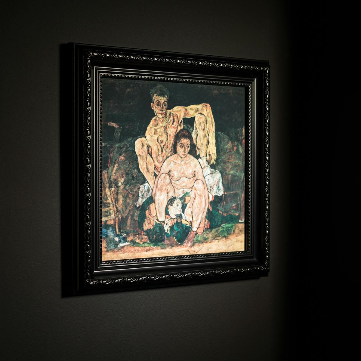 Photograph black ornate picture frame hung on a black wall. Inside the frame is a print of an oil painting by Egon Schiele titled, 'The Family'. It depicts Schiele himself at the far back, his sinewy nude body hunched behind his wife, Edith, who looks off to the side, while a child is curled between her feet.