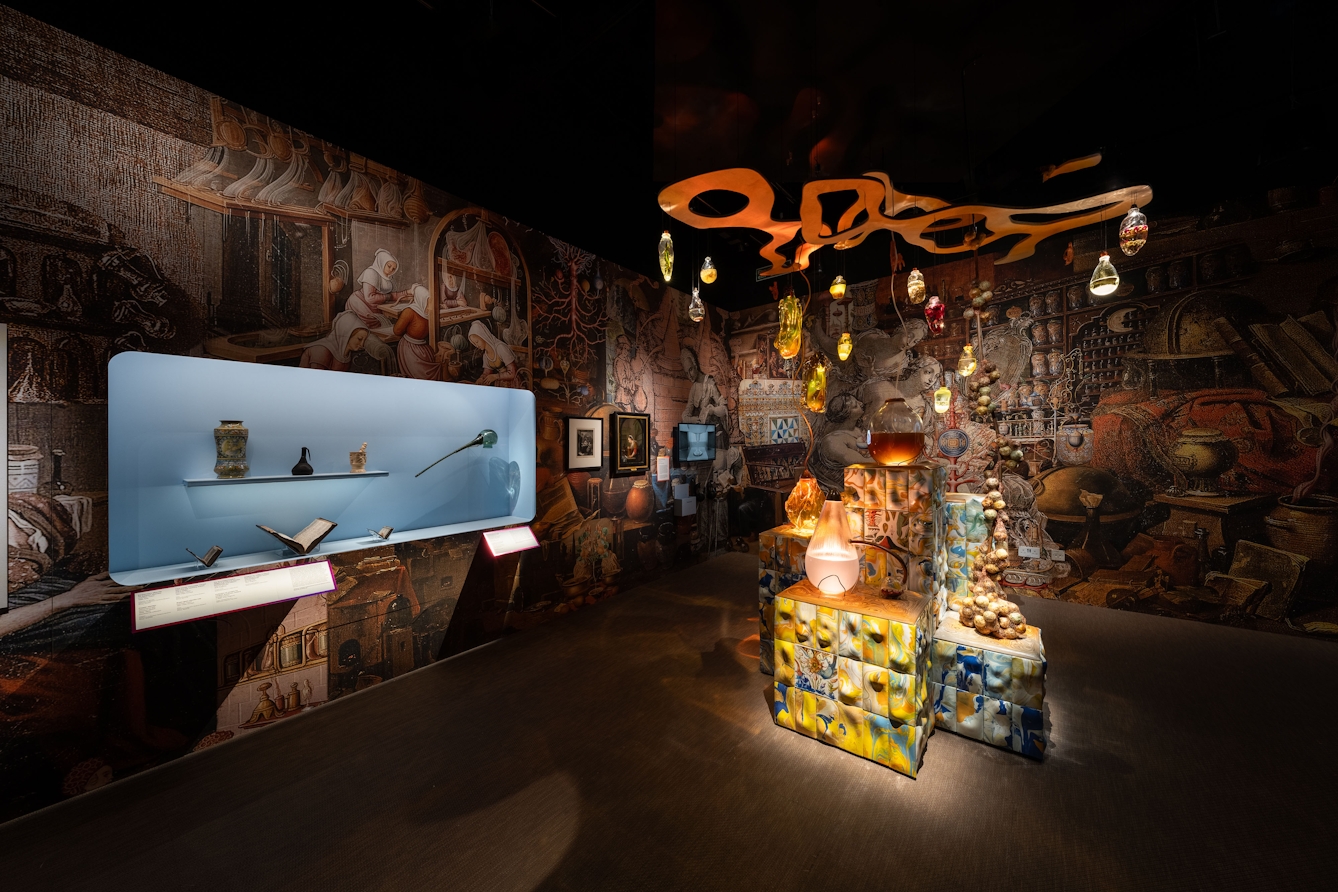 An exhibition space called 'Beauty Sensorium' containing several large glass vessles filled with liquid and other objects on decorated plinths in the central space with small bottles of liquid suspended on a mobile above them. The walls are covered in overlapping enlarged images of historical artworks and a glass-fronted display case containing several historical artefacts