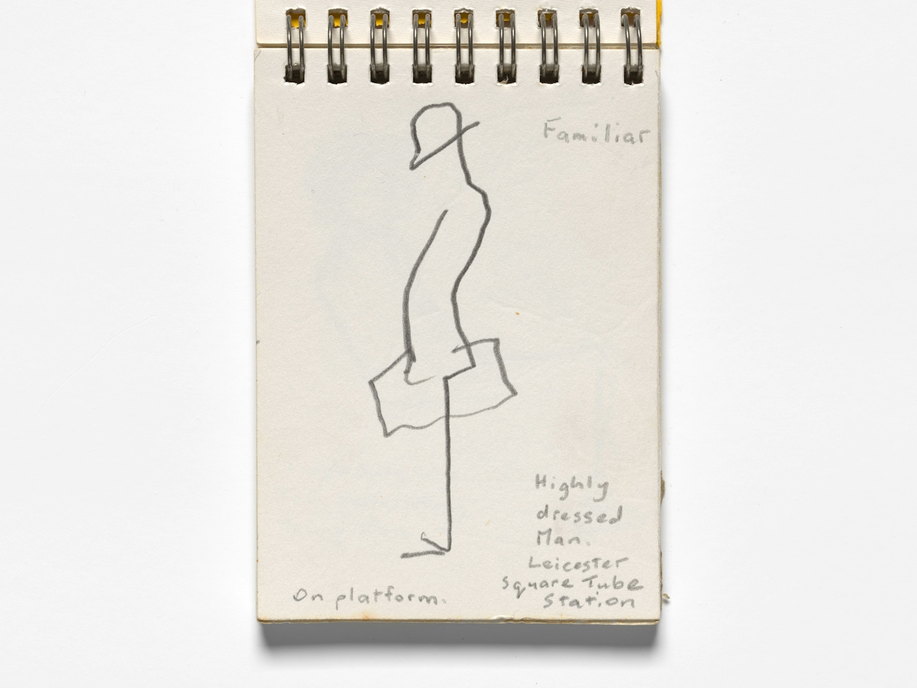 Photograph of an open sketchbook on a white background.  A simple line sketch is visible, showing a man in a hat, carrying a case, stood on a station platform. Although only made up of a few lines, the sketch conveys a sense of waiting and contemplative thought. The sketch is hand annotated with the words, 'Familiar, Highly dressed man. Leicester Square Tube Station. On platform."