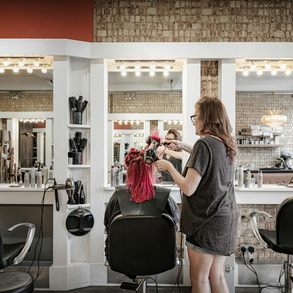 Photograph showing a woman from behind sitting in a hairdressing salon chair, wearing a rubber hairdressing collar. To her right the hairdresser is blow drying her pink hair. In front of her are three mirrors, lit with 4 bare bulbs per mirror. Behind the mirror the walls are made up of bare bricks.